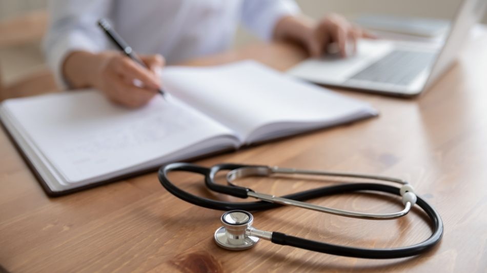 Stethoscope on wooden desk beside open notebook and laptop, with hand writing notes