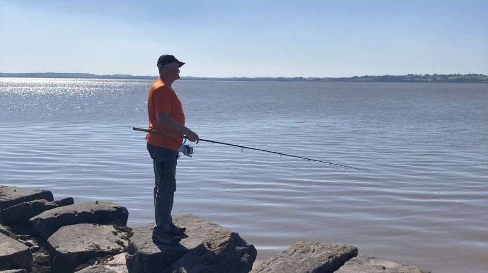 Person fishing from rocky shoreline, wearing orange shirt, overlooking calm sunlit water.