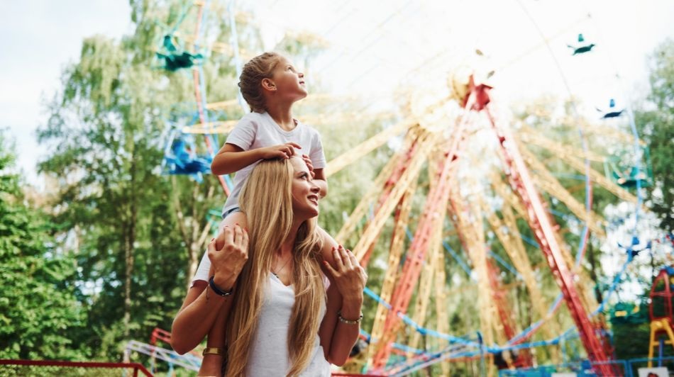 Child sitting on an adult’s shoulders at a colourful amusement park with spinning rides in the background.