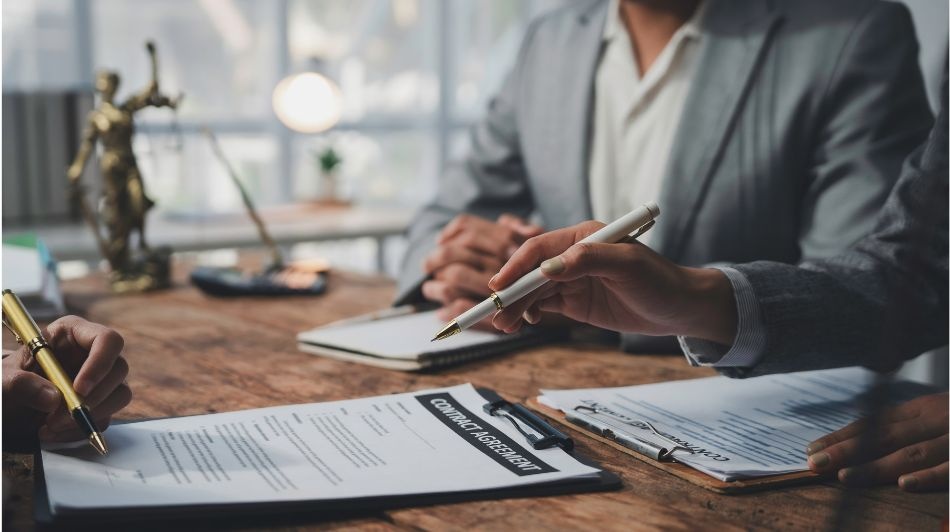 Legal professionals reviewing and signing a contract agreement at a conference table with legal documents and a scales of justice statue in the background.