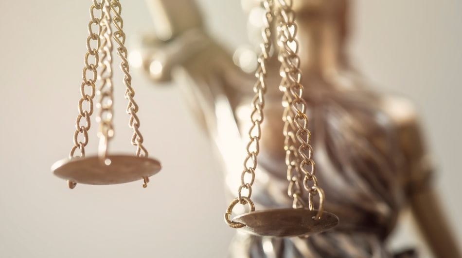 Close-up of golden scales of justice hanging from chains, with a blurred Lady Justice statue in the background.