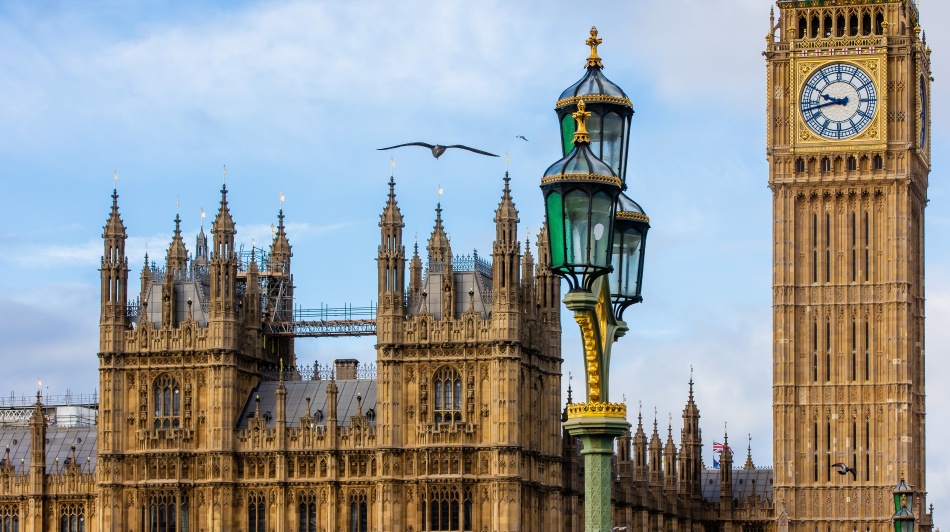 The Houses of Parliament and Big Ben in London beneath a cloudy sky, with an ornate Victorian street lamp and birds flying overhead.