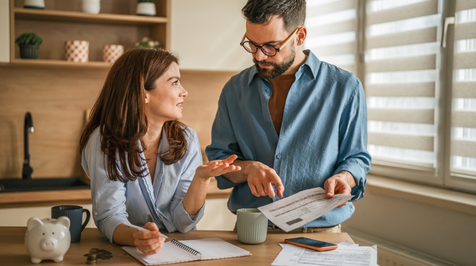 Two people reviewing household bills and budgeting documents at a kitchen table with a piggy bank, notebook, and coins.