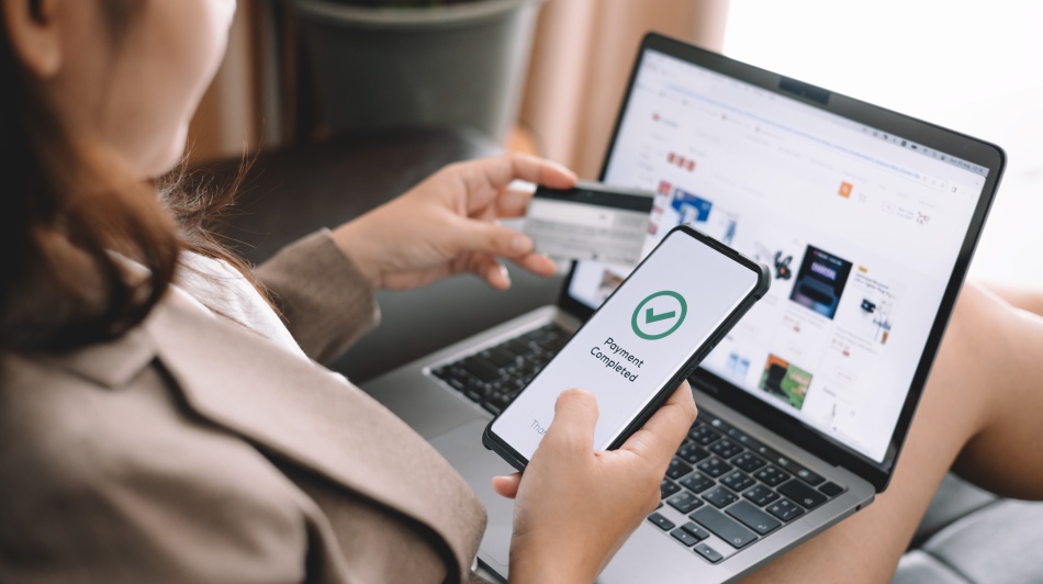 Person holding a credit card and smartphone displaying “Payment Completed” while shopping online on a laptop.