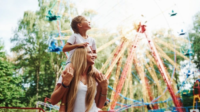 Child sitting on an adult’s shoulders at a colourful amusement park with spinning rides in the background.