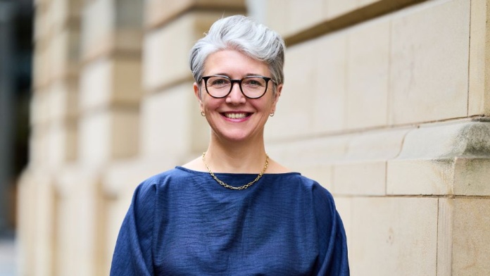 Professional headshot of a person with short grey hair and glasses, standing outdoors against a stone building.