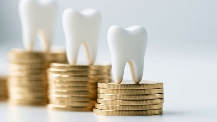 Three white tooth models displayed on stacks of gold coins, representing dental funding, costs, or investment in oral healthcare.