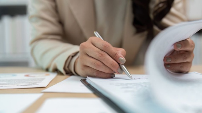 Close-up of a person reviewing documents and signing paperwork with a silver pen at a desk.