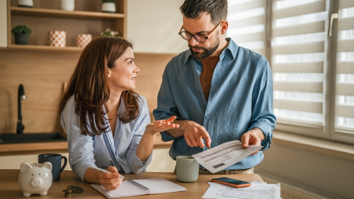 Two people reviewing household bills and budgeting documents at a kitchen table with a piggy bank, notebook, and coins.