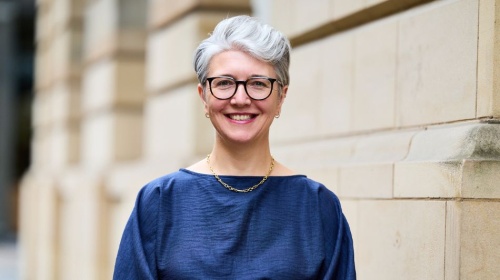 Professional headshot of a person with short grey hair and glasses, standing outdoors against a stone building.
