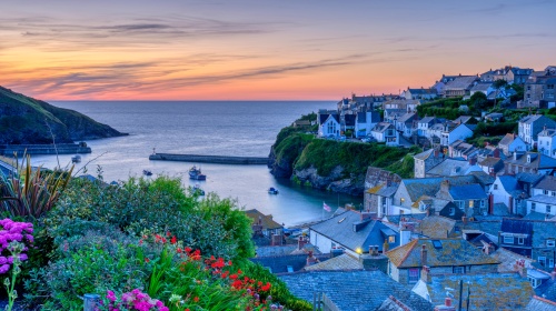 Colourful coastal village at sunset with harbour, boats and flower-lined hillside.