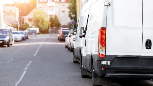 White delivery van parked along a suburban street lined with cars and residential buildings in daylight.