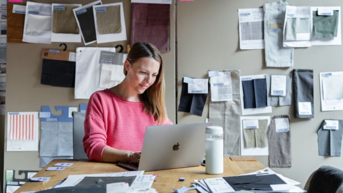 Person working on a laptop at a desk surrounded by fabric swatches and textile samples pinned to a studio wall.