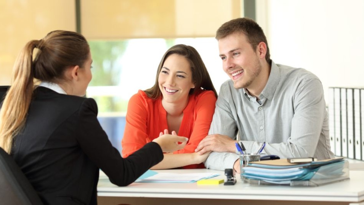 Person in business attire meeting with two clients across a desk during a consultation in a modern office.