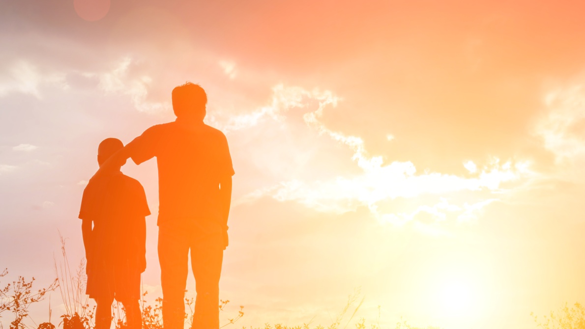 Silhouetted adult and child standing together on a grassy hill at sunset, illuminated by warm golden light and dramatic clouds.