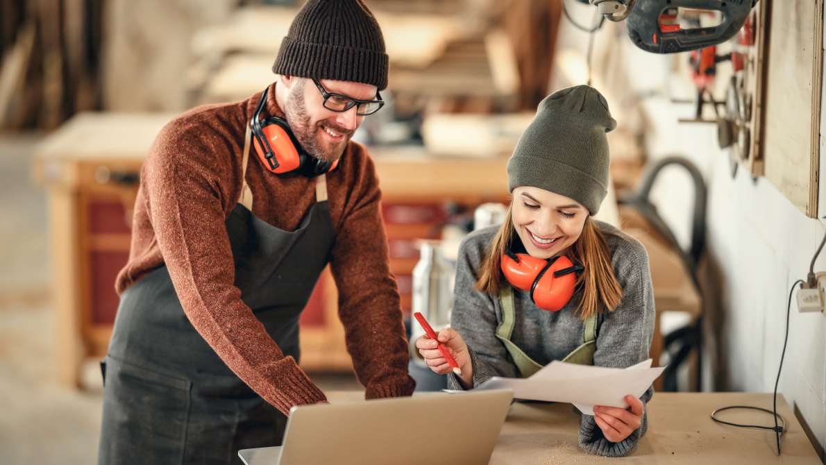 Two woodworkers in a workshop reviewing plans beside a laptop, wearing aprons and hearing protection near woodworking equipment.