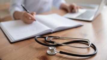 Stethoscope on wooden desk beside open notebook and laptop, with hand writing notes