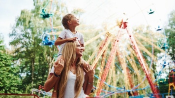 Child sitting on an adult’s shoulders at a colourful amusement park with spinning rides in the background.