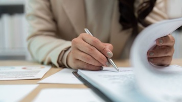Close-up of a person reviewing documents and signing paperwork with a silver pen at a desk.