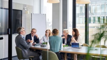 Modern office boardroom meeting with professionals gathered around a conference table and presentation easel.
