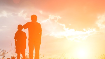 Silhouetted adult and child standing together on a grassy hill at sunset, illuminated by warm golden light and dramatic clouds.