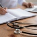 Stethoscope on wooden desk beside open notebook and laptop, with hand writing notes