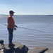 Person fishing from rocky shoreline, wearing orange shirt, overlooking calm sunlit water.