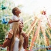 Child sitting on an adult’s shoulders at a colourful amusement park with spinning rides in the background.
