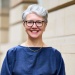 Professional headshot of a person with short grey hair and glasses, standing outdoors against a stone building.
