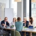 Modern office boardroom meeting with professionals gathered around a conference table and presentation easel.