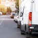 White delivery van parked along a suburban street lined with cars and residential buildings in daylight.