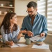 Two people reviewing household bills and budgeting documents at a kitchen table with a piggy bank, notebook, and coins.