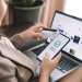 Person holding a credit card and smartphone displaying “Payment Completed” while shopping online on a laptop.