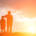 Silhouetted adult and child standing together on a grassy hill at sunset, illuminated by warm golden light and dramatic clouds.