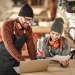 Two woodworkers in a workshop reviewing plans beside a laptop, wearing aprons and hearing protection near woodworking equipment.